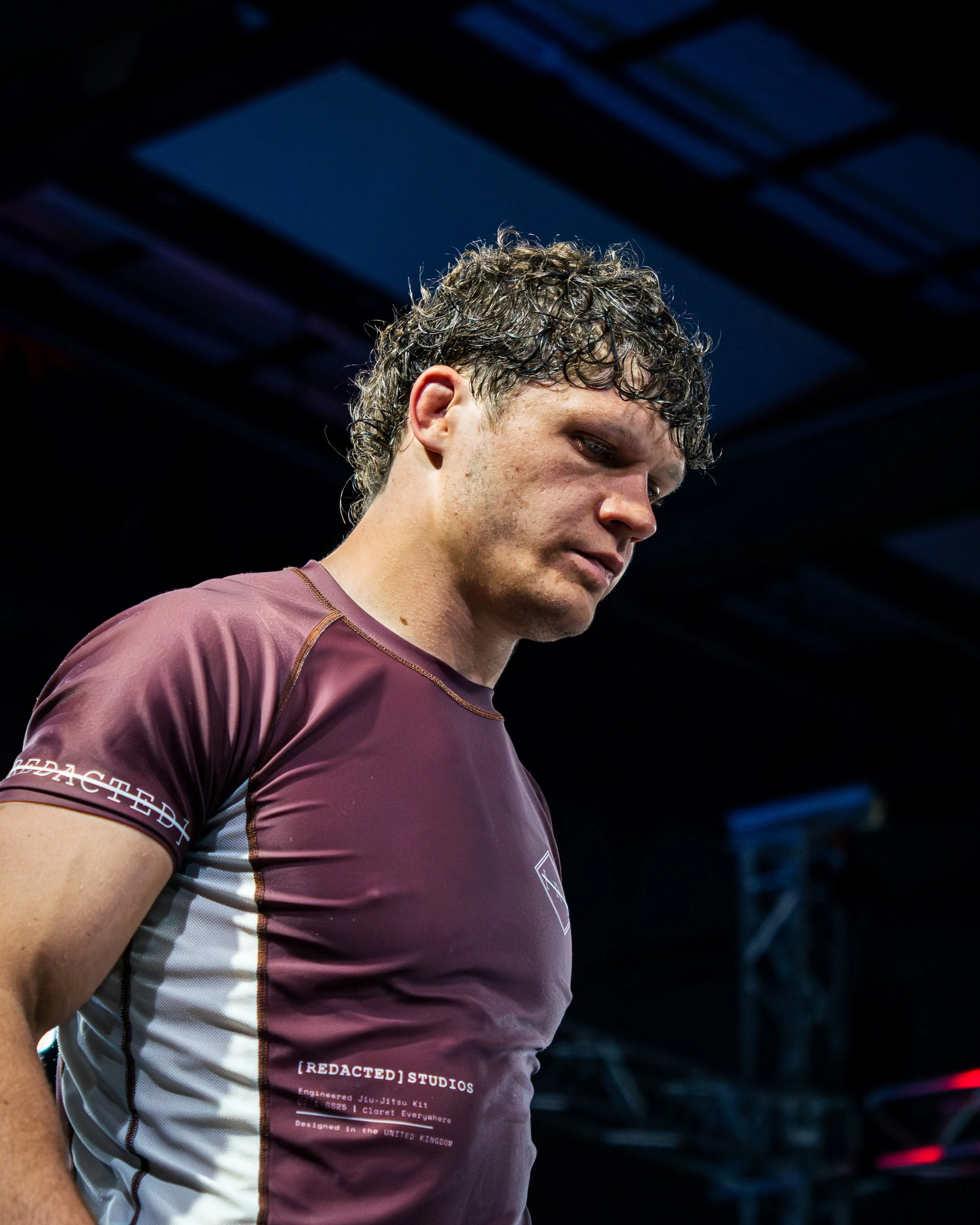 A young man with curly brown hair and light skin looks downward, wearing a maroon and white sports jersey with black text, in a dark indoor setting with blue light fixtures overhead.
