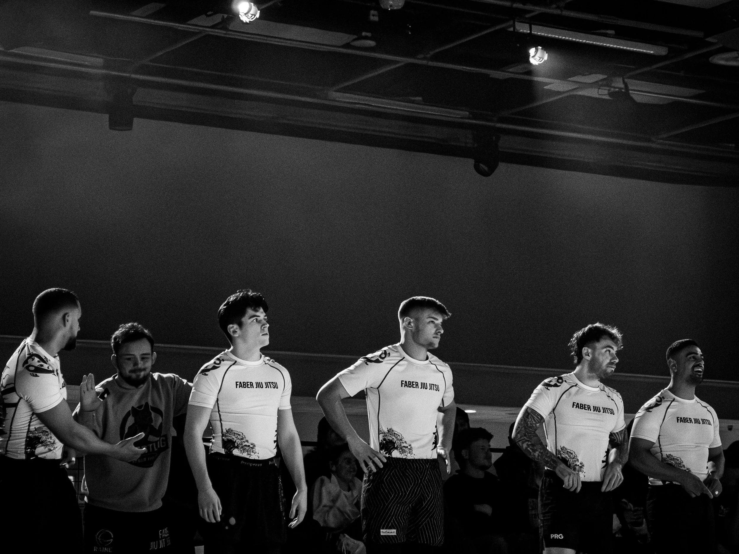 A black and white photograph of a group of young men standing in a line, wearing sports jerseys with the text "Faber Jiu Jitsu" on them. They appear to be at an indoor event, possibly a sports competition or team gathering, with some holding bouquets