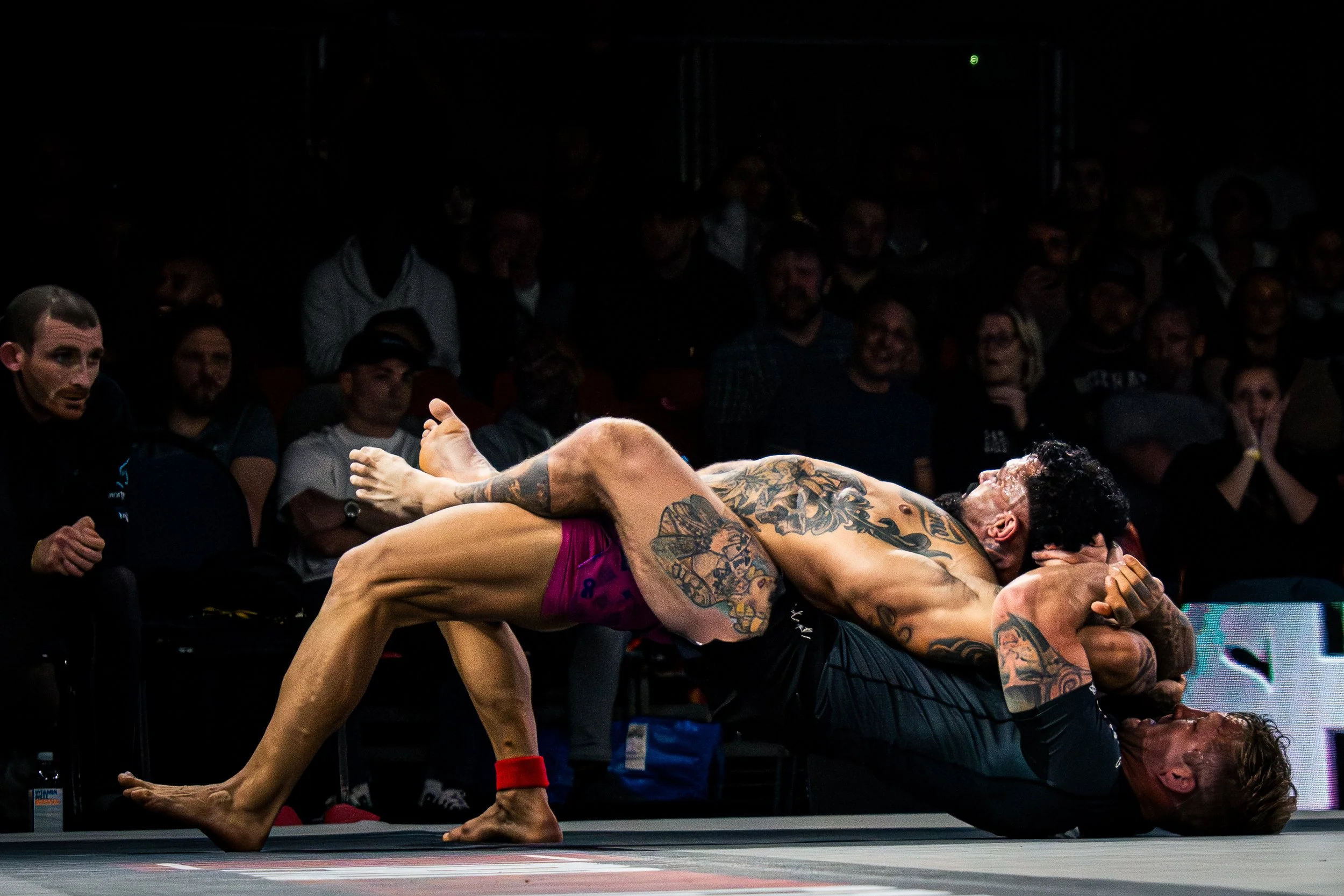 Two wrestlers engaged in a match, with one wrestler in a dominant position over the other, in a wrestling ring surrounded by spectators.