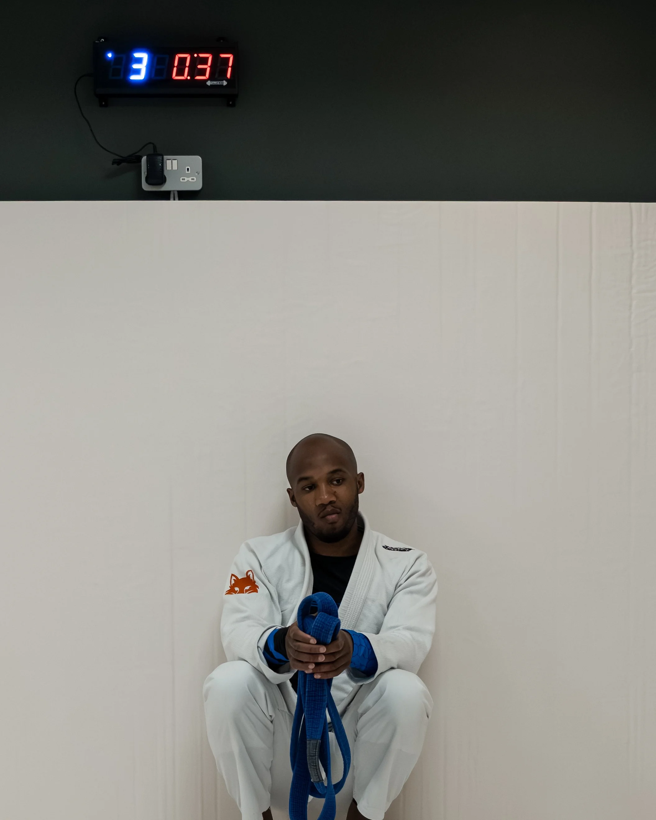 A man wearing a white martial arts gi with blue gloves, sitting on a white floor, holding a blue belt, in front of a plain white wall and a black wall clock displaying the time and temperature.