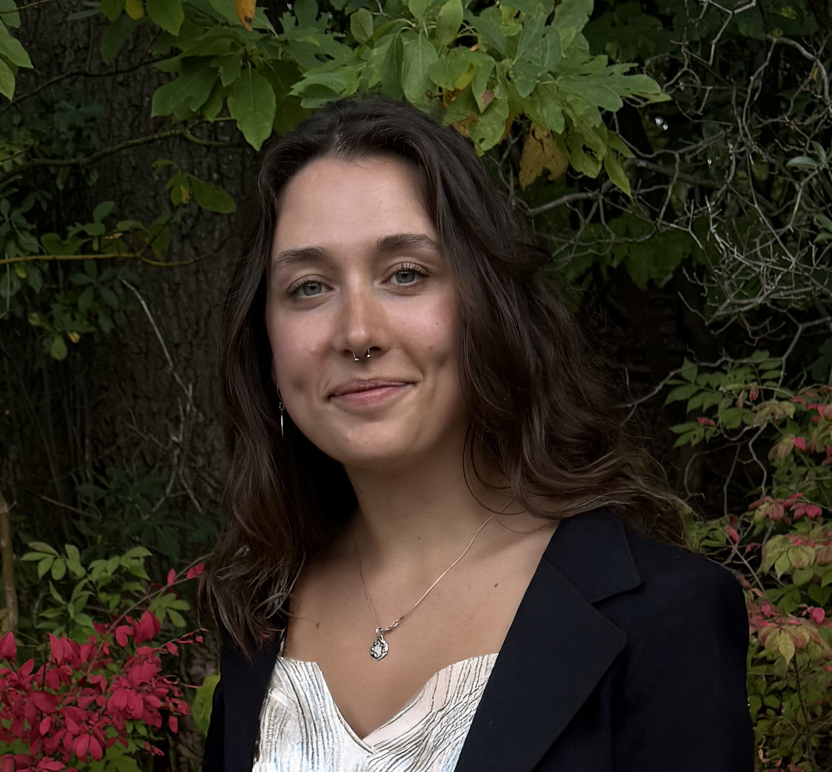 A woman with wavy brown hair, wearing a black blazer and a white top with a necklace, standing outdoors in front of green foliage with some pink and yellow leaves.