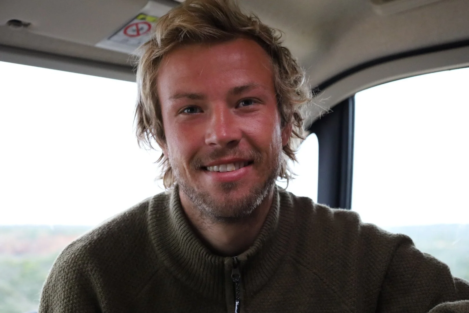 Et bilde av en smilende mann med lyse, krøllete hår, som sitter inne i et kjøretøy med store vinduer, antagelig i en natur. Han har på seg en grønn jakke med glidelås.