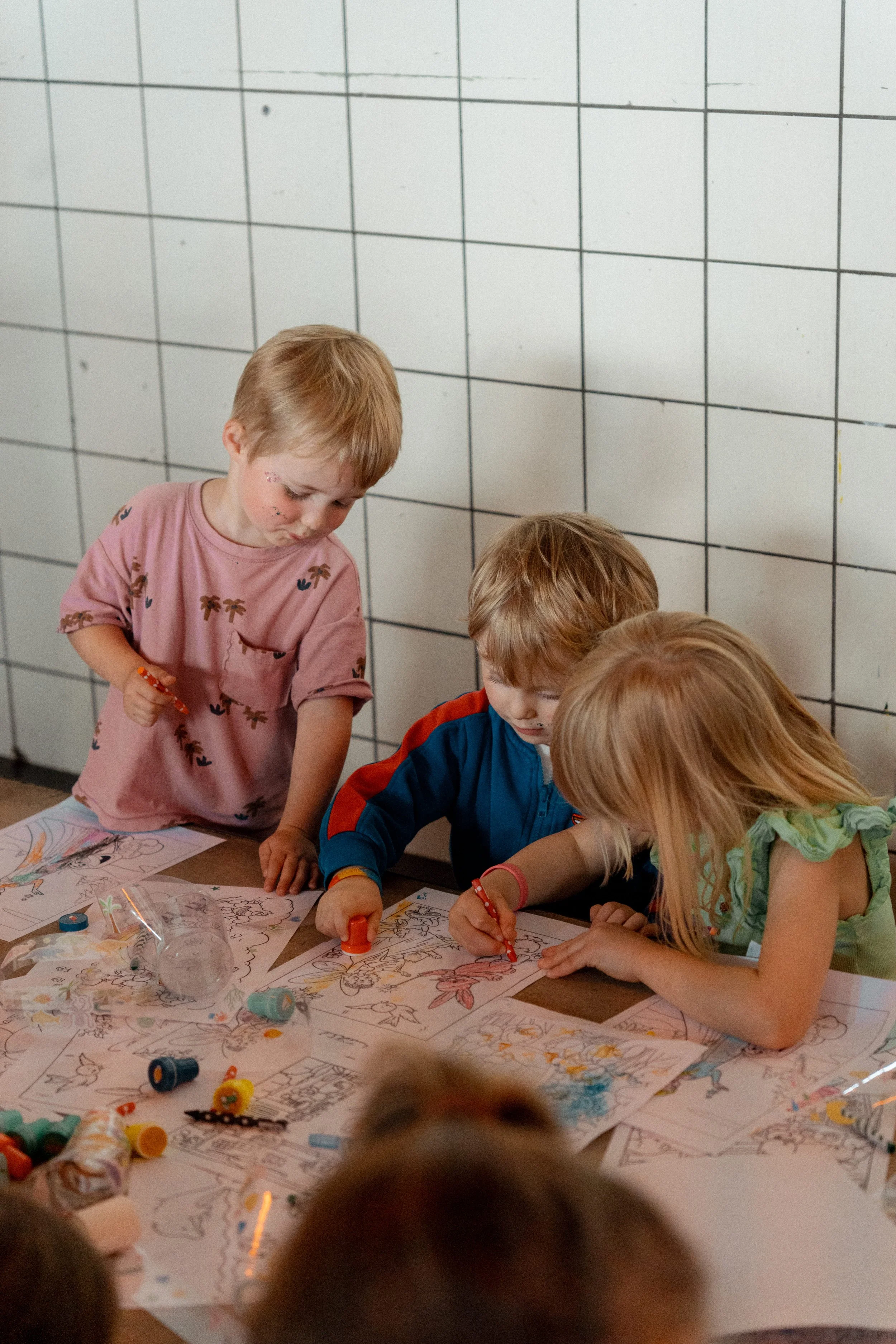Several children are gathered around a table, coloring and decorating large sheets of paper with various drawings and designs, with some crayons and decorations scattered on the table. A tiled wall is visible in the background.