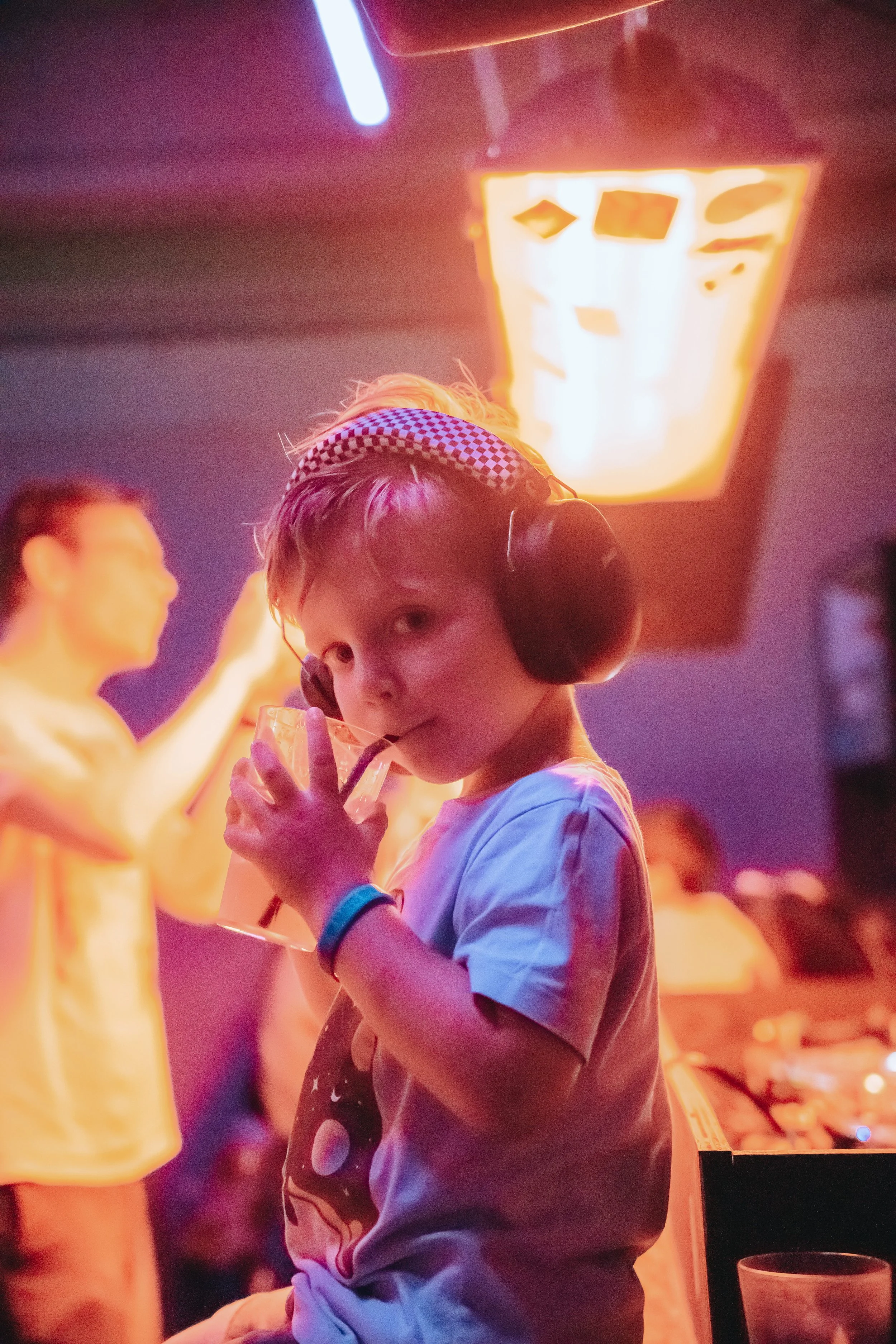 Young boy with headphones drinking from a glass at a social gathering or party with blurred people in the background, illuminated by warm light and neon sign.