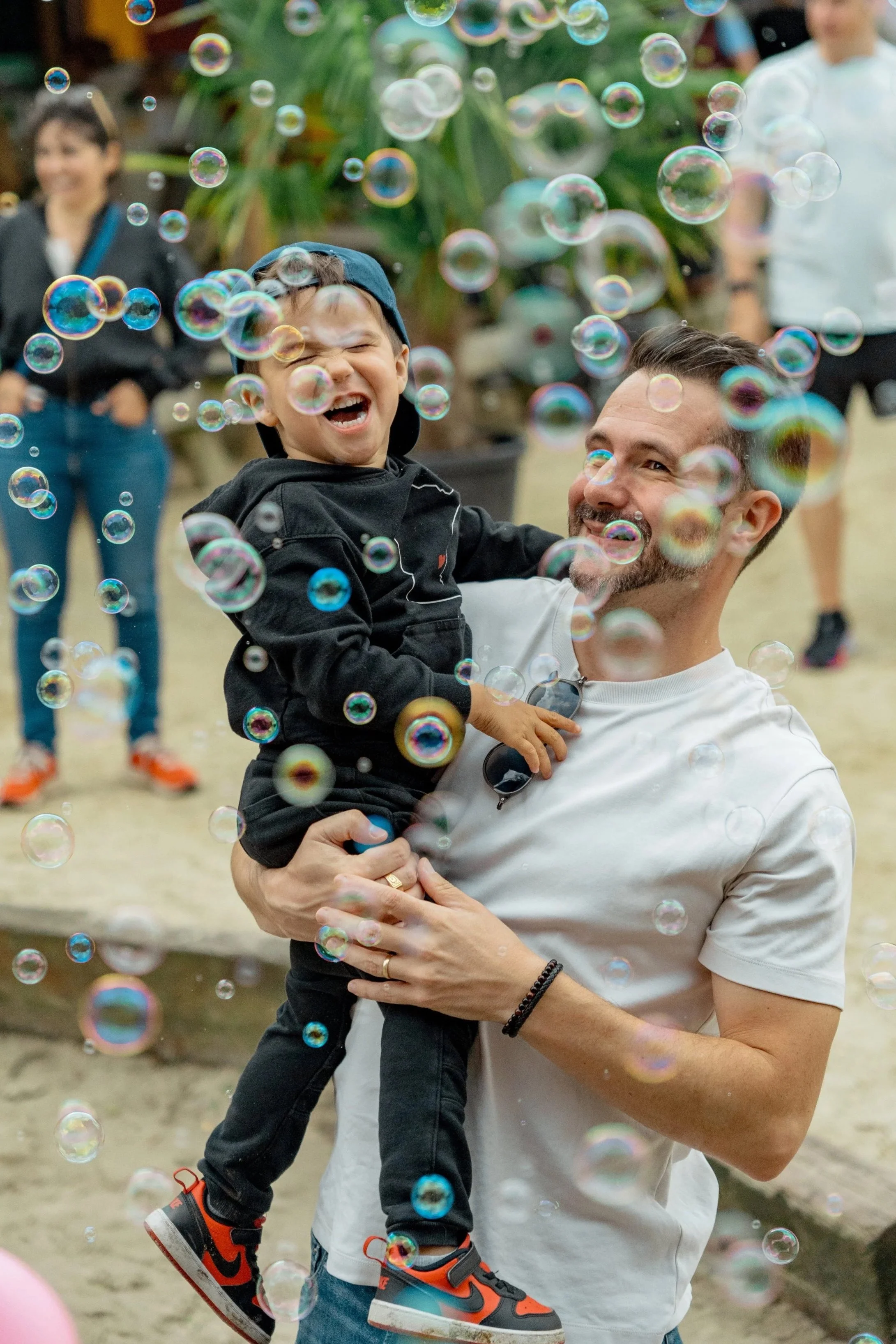 A man holding a smiling young boy surrounded by floating bubbles at an outdoor gathering.