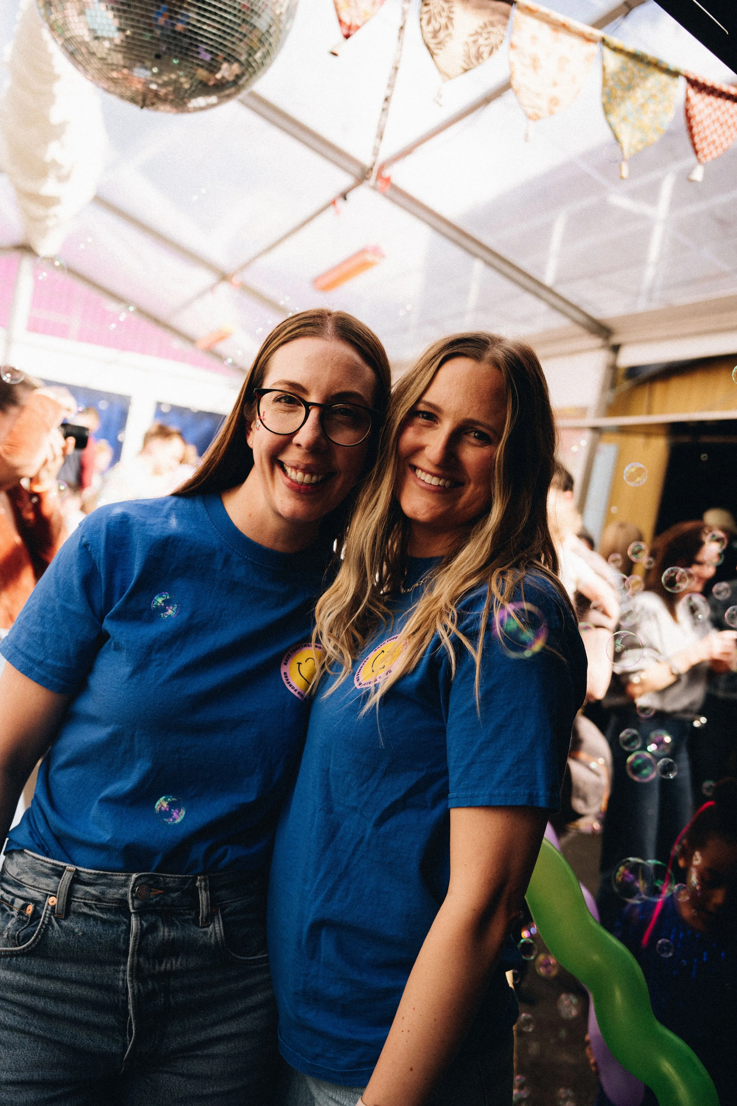 Two women smiling, wearing matching blue shirts, at a festive indoor event decorated with bunting, bubbles, and colorful balloons.