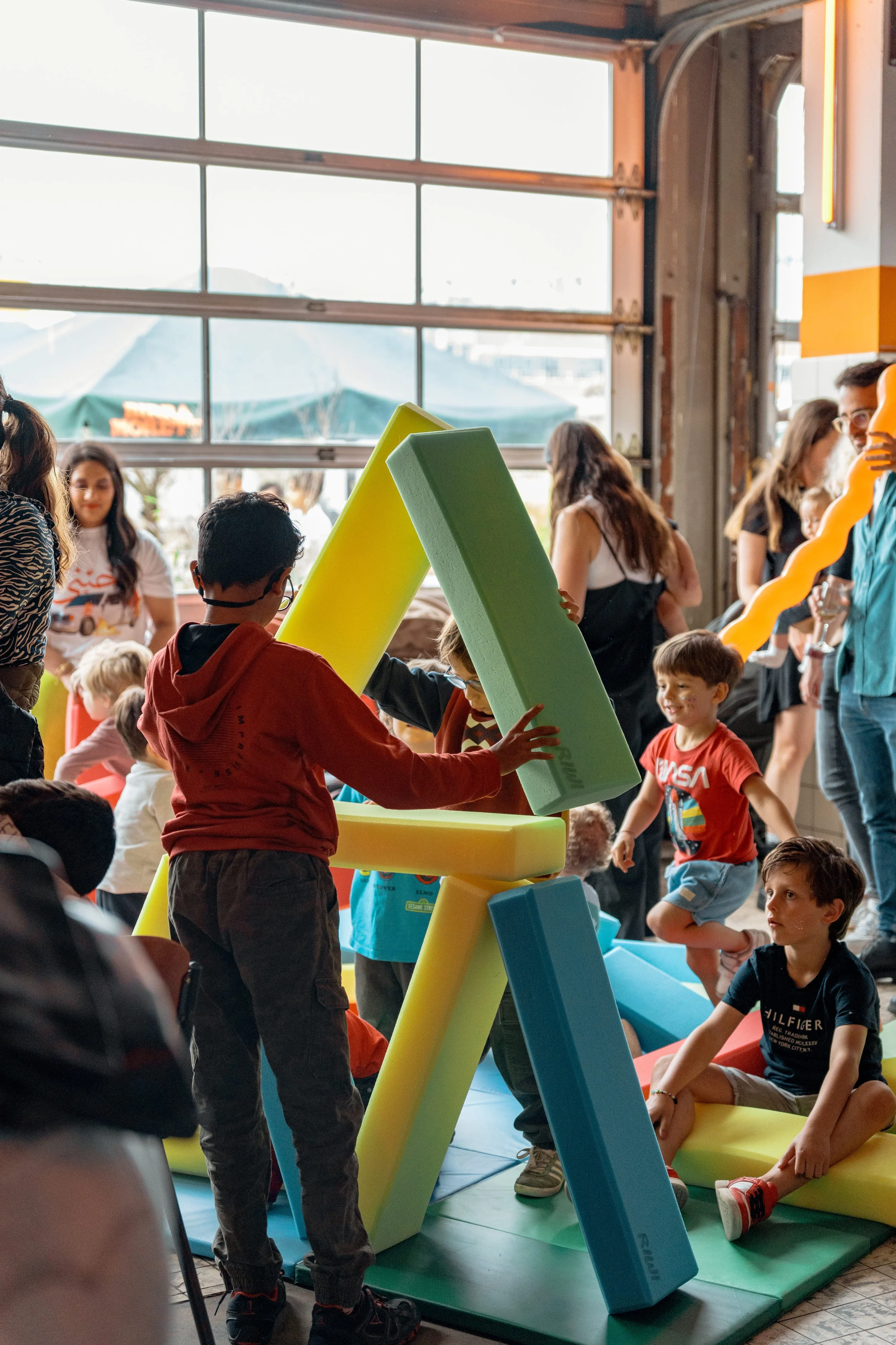 Children and adults gathered inside a colorful play area with foam blocks for stacking and building, near a large garage door with natural light coming through.