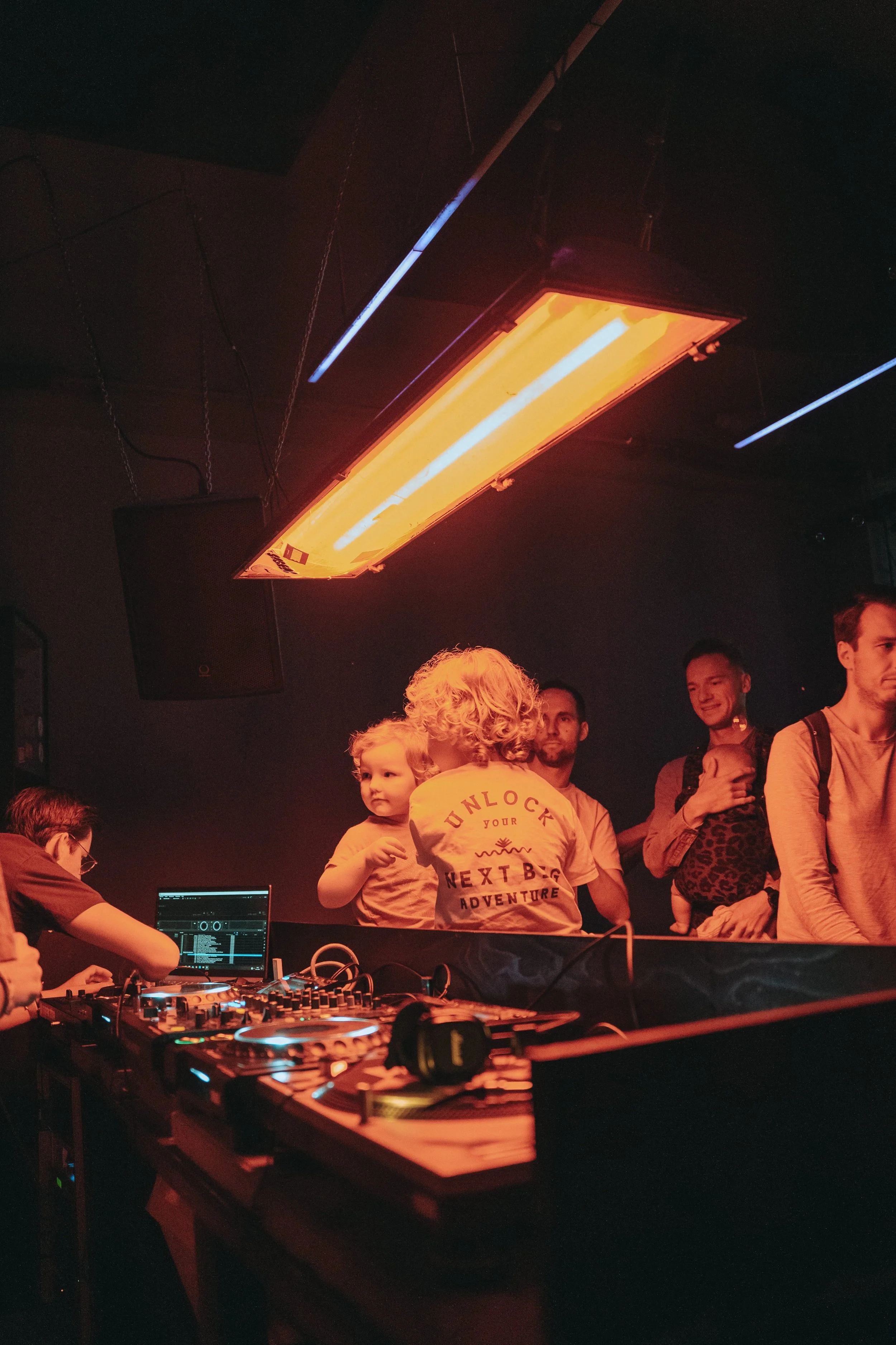 A DJ booth with a mixing console and a laptop, surrounded by a group of people, including a woman with curly blonde hair holding a child, in a dimly lit venue with a large orange lighting fixture overhead and a speaker on the ceiling, creating a lively atmosphere.