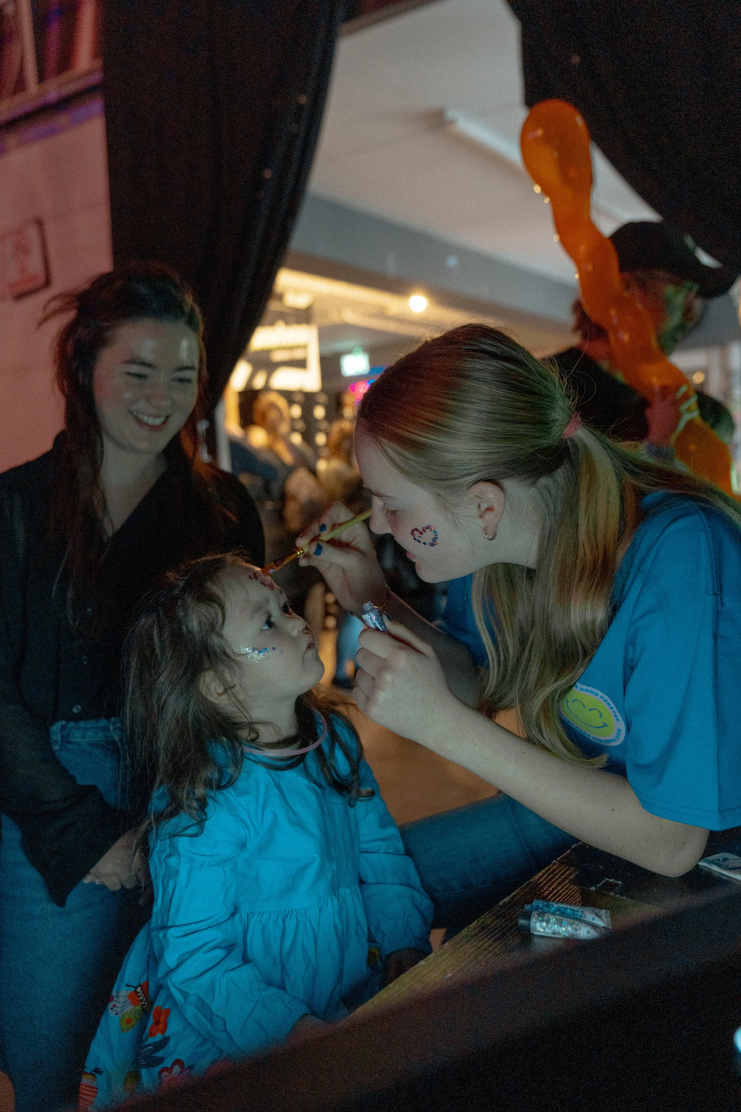 A woman is painting a child's face at an indoor event, while another woman laughs nearby and a crowd is visible in the background.