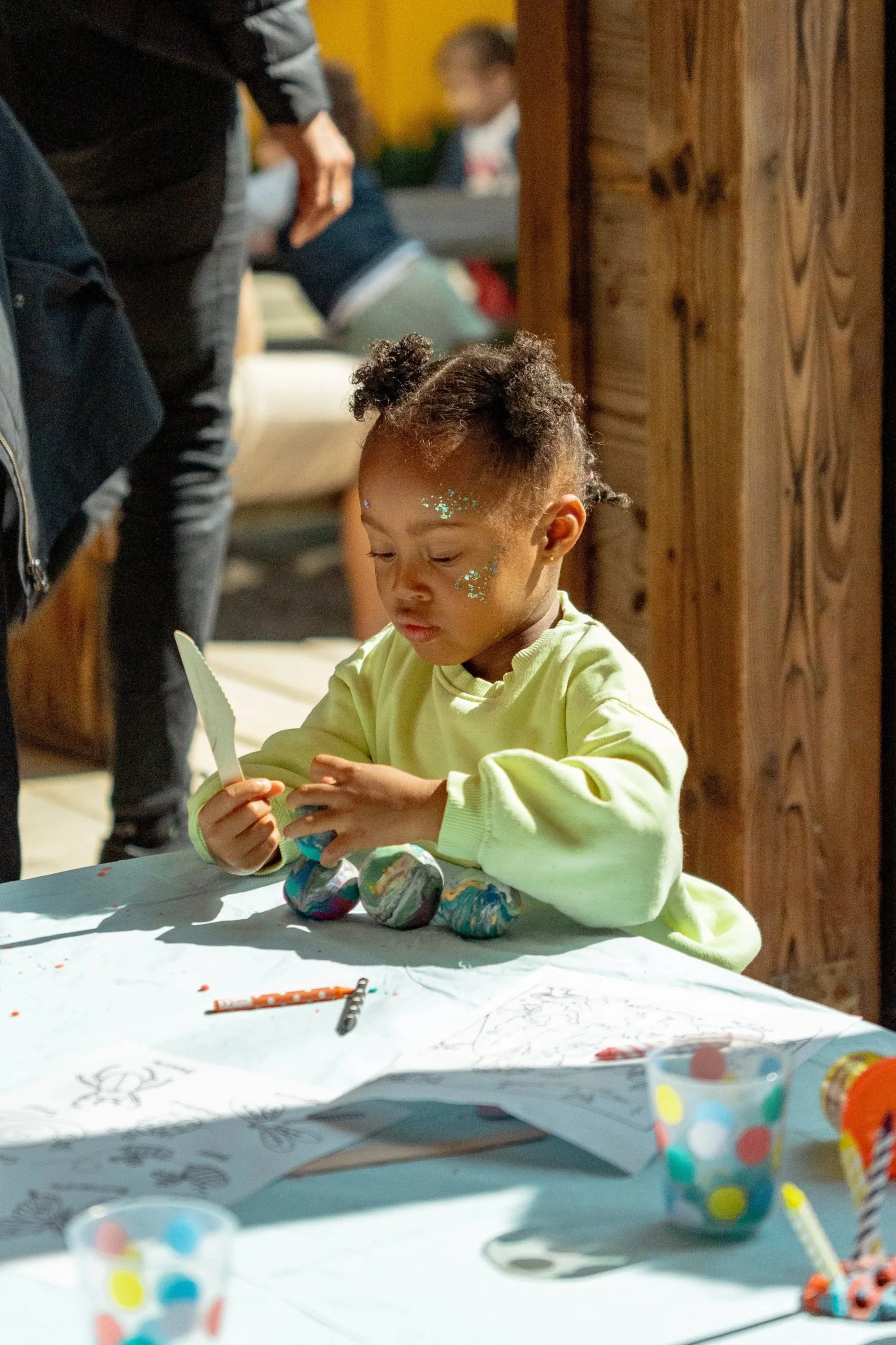 A young girl with dark, curly hair in puffs and glitter on her face is sitting at a table, participating in a craft activity involving painted rocks. The table is covered with papers, markers, and cups of paint. Other people are visible in the blurry background.