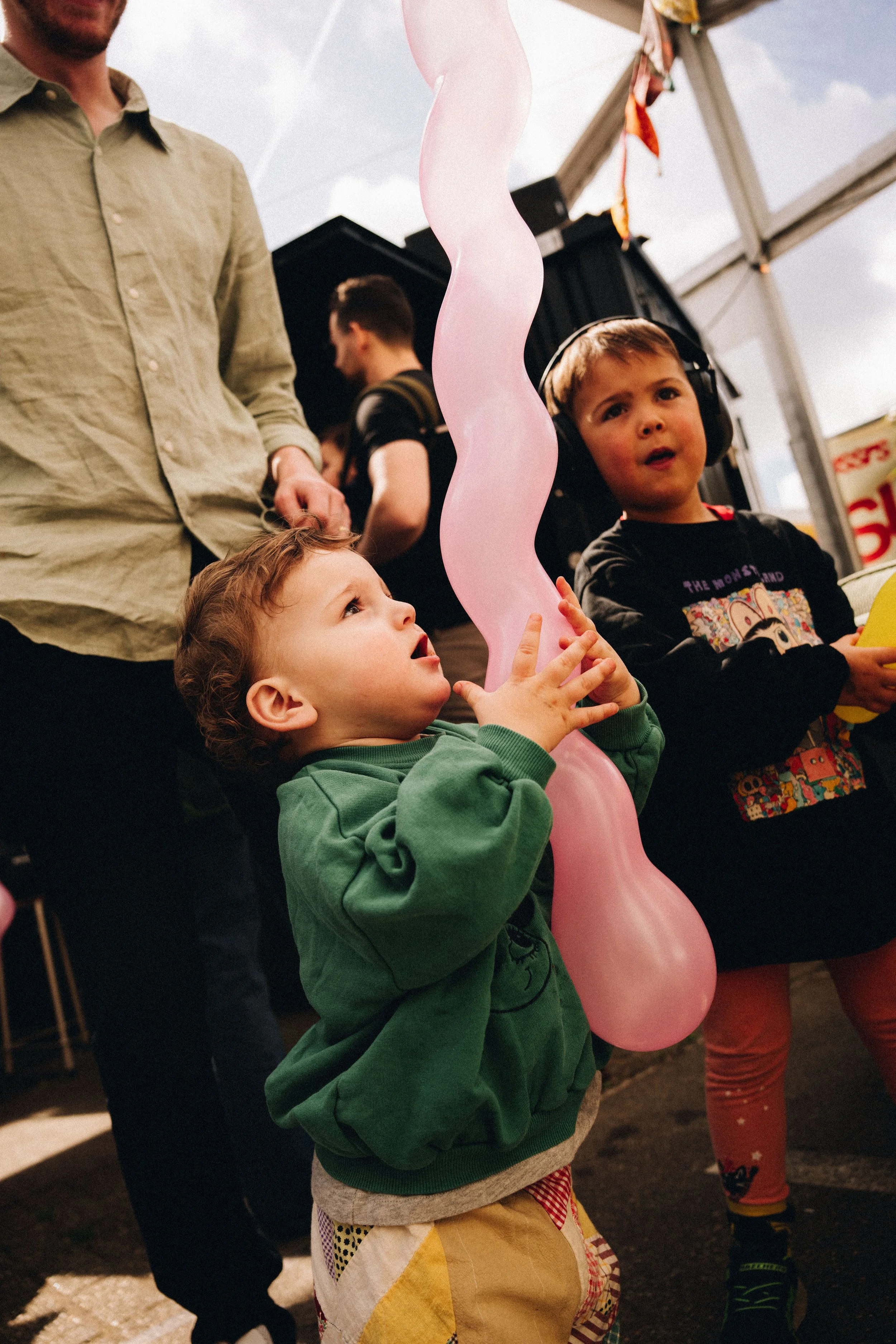 Two young children, a boy and a girl, are at an outdoor event under a large tent. The boy with a green hoodie is blowing up a large pink balloon, while the girl with headphones and a black shirt is watching him. Other people are seen in the background.