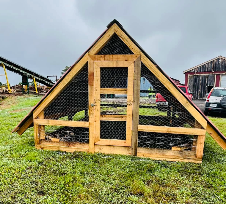 A wooden chicken coop with a triangular roof, covered with wire mesh, situated on a grassy area.