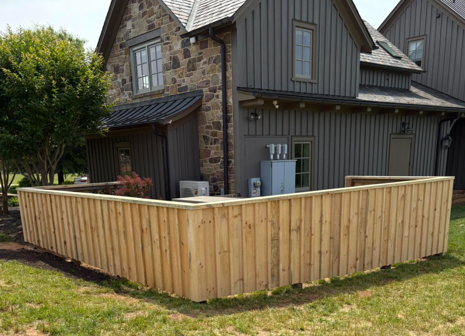 A new wooden fence surrounds a landscaped backyard adjacent to a modern house with dark gray siding and stone accents, with some trees and shrubs visible.