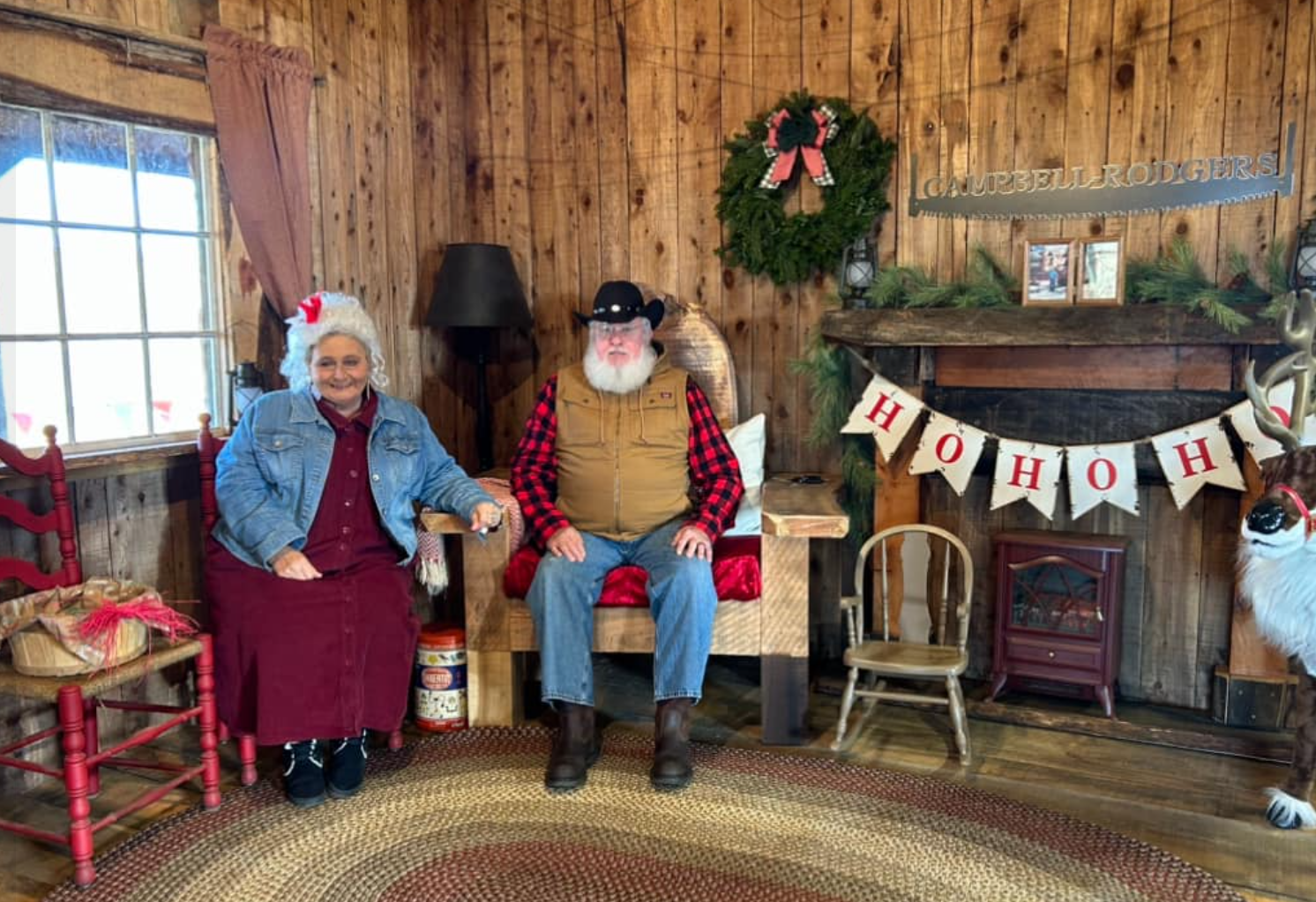 A woman and a man in cowboy attire sitting in a Christmas decorated room with wooden walls. The woman is smiling, wearing a red dress, a blue denim jacket, and a Santa hat. The man is dressed in a plaid shirt, a tan vest, jeans, and a black cowboy hat. The room has a Christmas wreath, a garland with