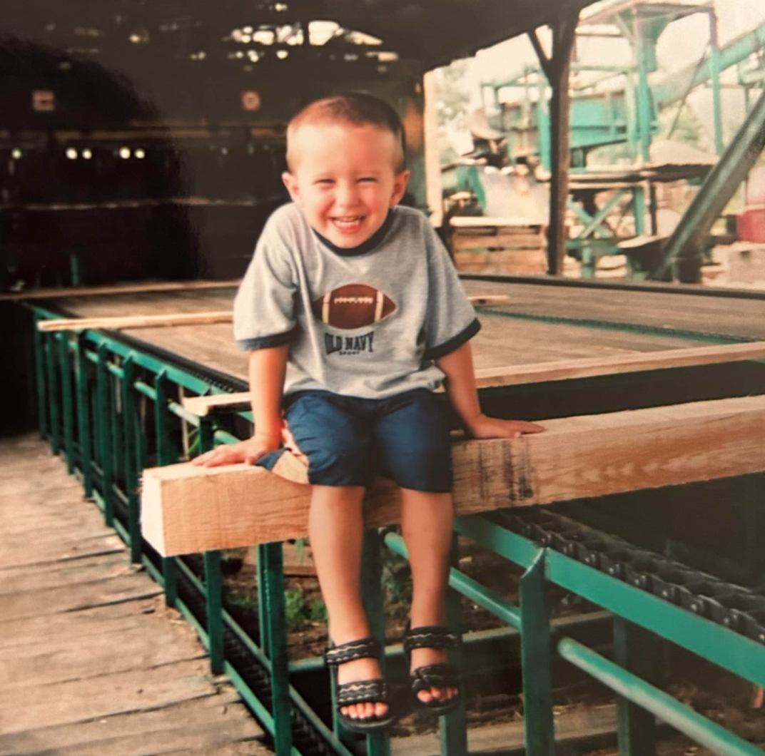 A young boy with short hair sitting on the edge of a wooden structure at an amusement park, smiling and wearing a gray T-shirt with a football graphic, dark shorts, and black sandals.