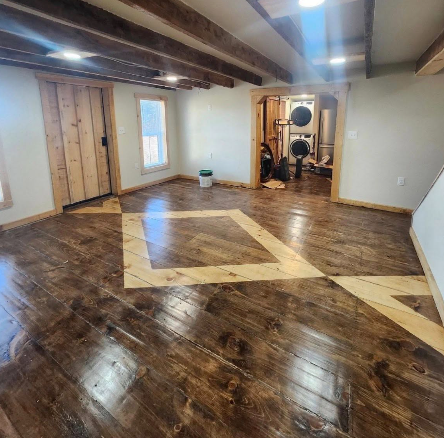 A room with hardwood floors, wooden beams on the ceiling, and a doorway leading to a laundry area with a washing machine and dryer. There are two windows, one with white trim, and a bucket on the floor.