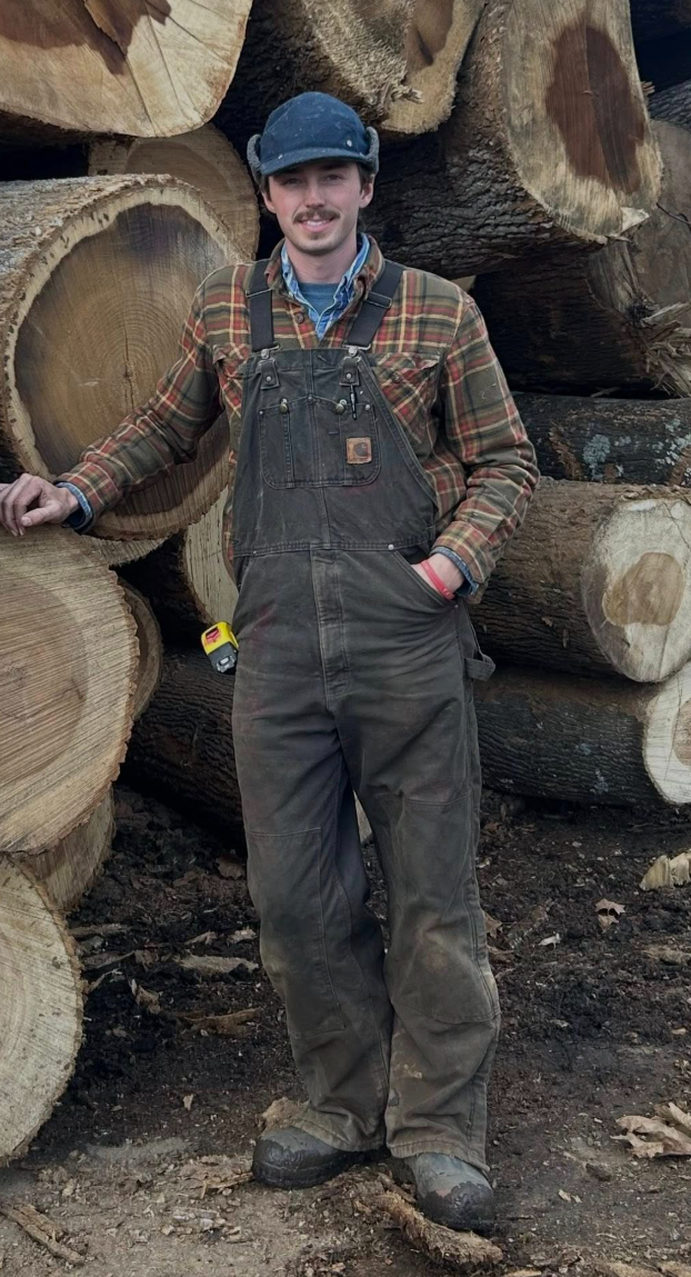 A man in work overalls and a baseball cap standing in front of cut logs, smiling, with one hand on a log and the other in his pocket.