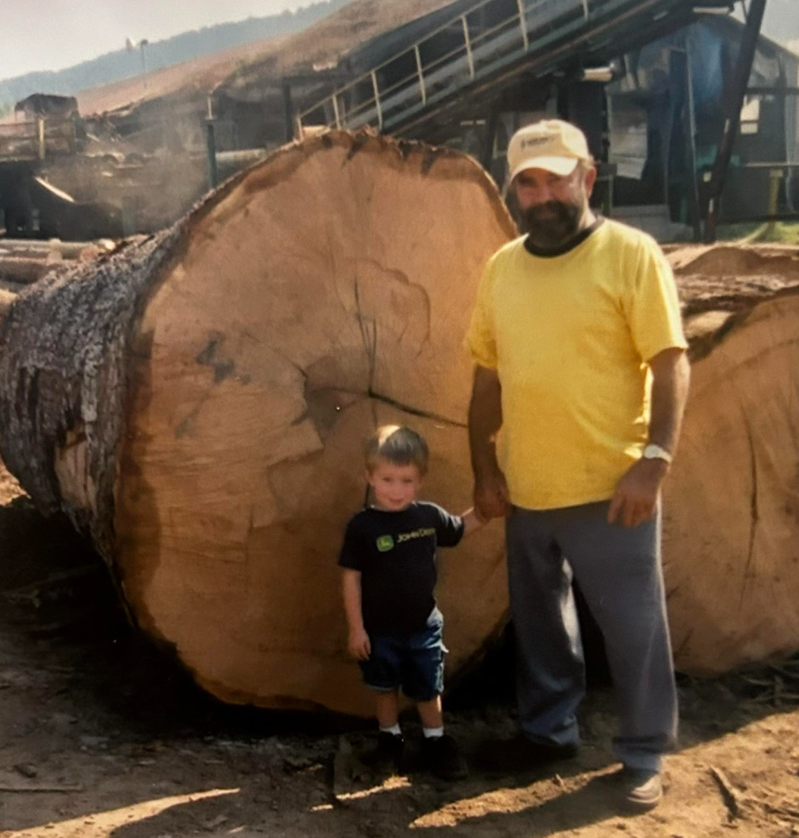 A man with a beard wearing a yellow T-shirt and a cap, standing next to a young boy in front of a large cut tree log at a lumber yard or sawmill. They are holding hands, surrounded by lumber and machinery.