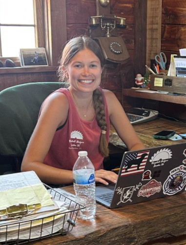 A young woman with long hair in a braid, wearing a red sleeveless top, sitting at a wooden desk with a laptop decorated with various stickers, a water bottle, and papers, inside a rustic wooden room.