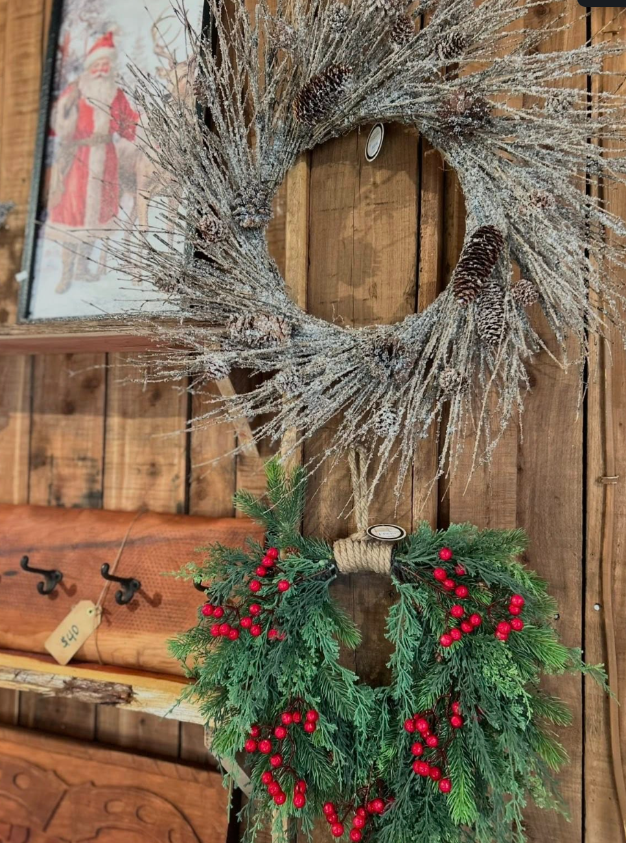 Decorative holiday wreaths hanging on a wooden wall. One wreath is made of frosted branches and pinecones, and the other is decorated with green foliage and red berries.