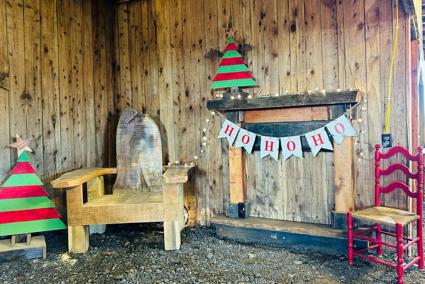 Rustic indoor Christmas scene with wooden walls, a wooden throne, and a red chair. Decorated with Christmas trees, string lights, and a banner with "HO HO HO".