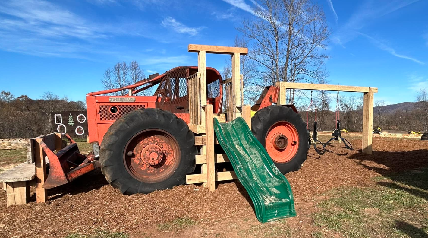 A playground made from a large red tractor with a green slide, set on wood chips with a clear blue sky and trees in the background.