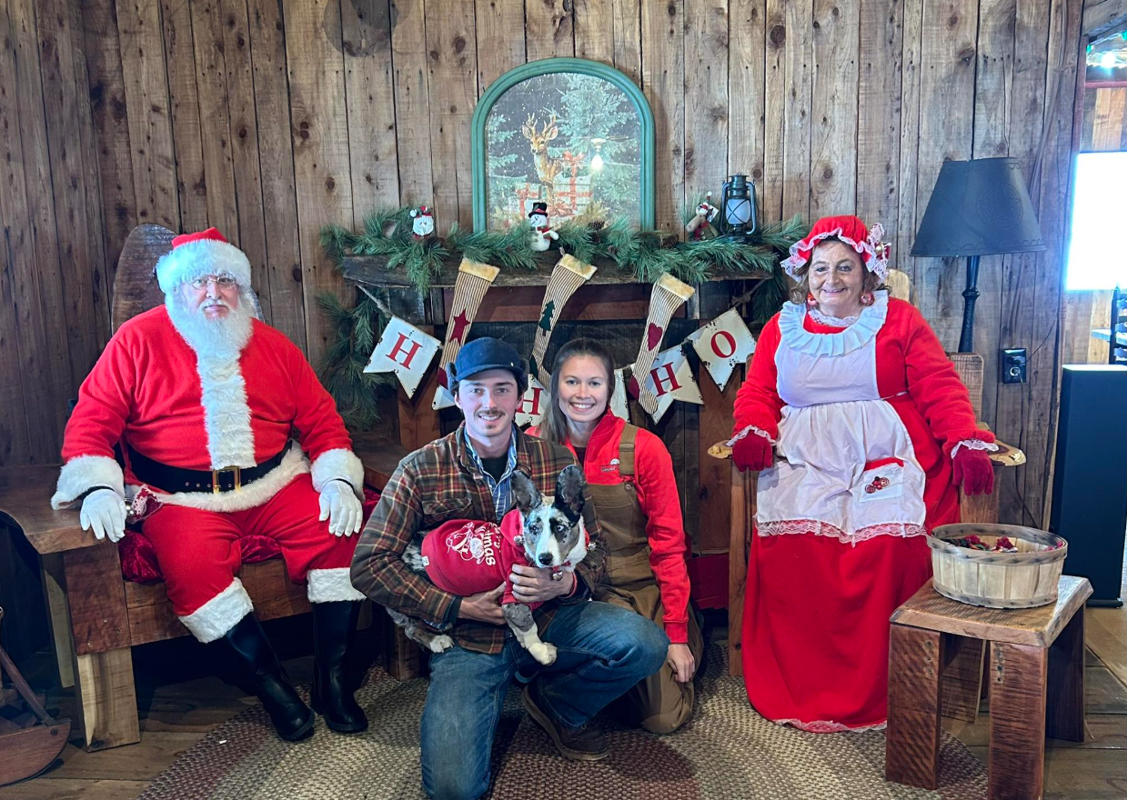 People dressed in Christmas costumes posing for a photo with a dog in front of a festive holiday backdrop.