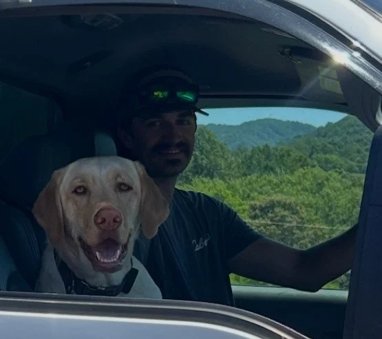Man driving a vehicle with a Labrador Retriever in the passenger seat, with lush green hills visible outside the window.