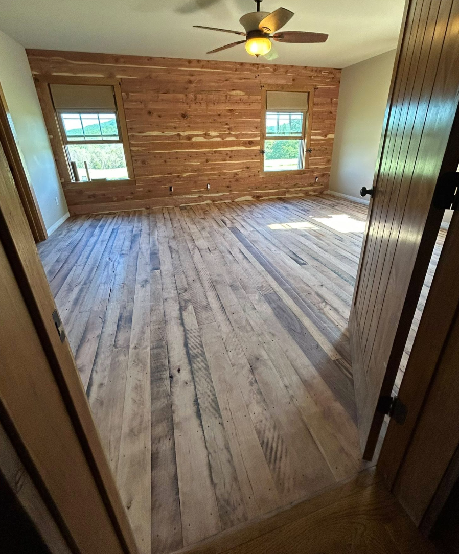 Empty room with wooden plank flooring, two windows, a ceiling fan, and a partially wood-paneled wall.