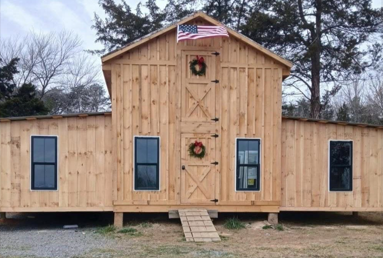 A large wooden barn with a central double door, decorated with two Christmas wreaths, and an American flag hanging above it. The barn has four black-framed windows and a small ramp leading up to the door. The background shows leafless trees and a cloudy sky.