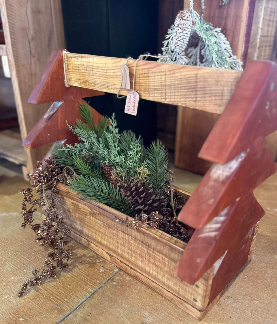 Wooden wheelbarrow filled with assorted pine branches, pinecones, and decorative winter greenery, placed on a wooden floor.