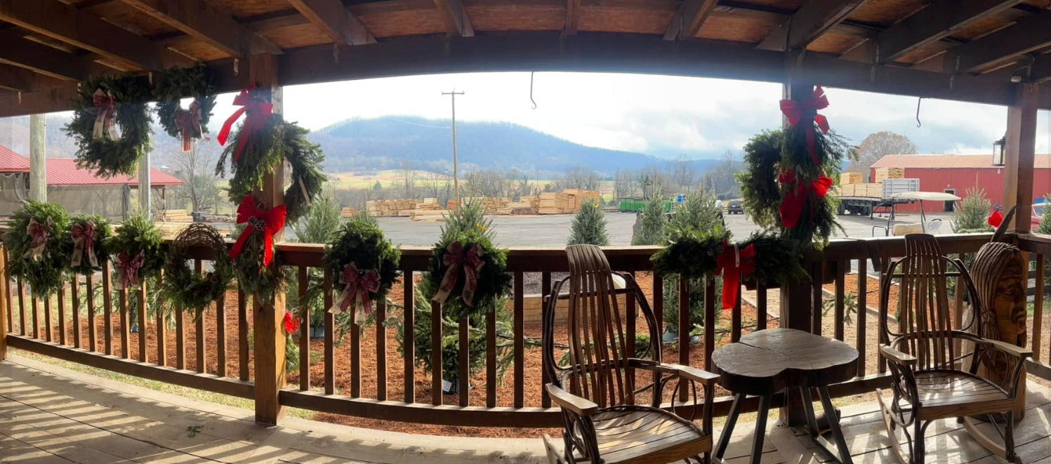 View from a covered porch decorated with Christmas wreaths and red bows, overlooking a farm area with trees, barns, and a mountain in the background.