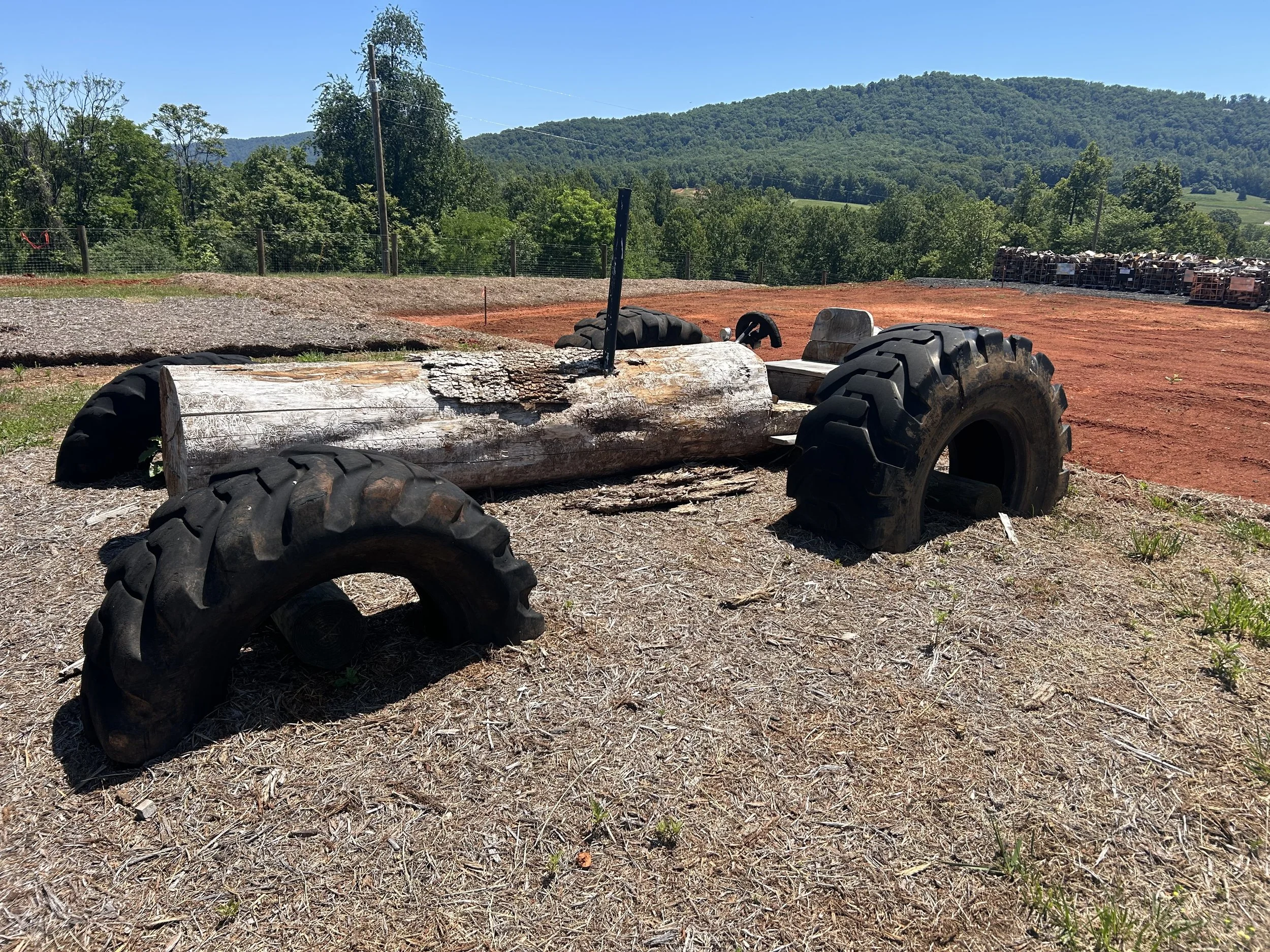 A rural outdoor scene with a tractor part, a log, and a mountain backdrop on a clear, sunny day.