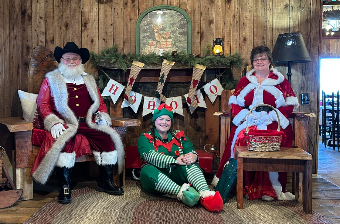 A festive holiday scene with two people dressed as Santa Claus and Mrs. Claus sitting on chairs, and a person dressed as an elf sitting on the floor between them, all smiling. The background features a wooden wall, a fireplace with Christmas stockings, and holiday decorations including a banner spelling 'HOHOHO'.