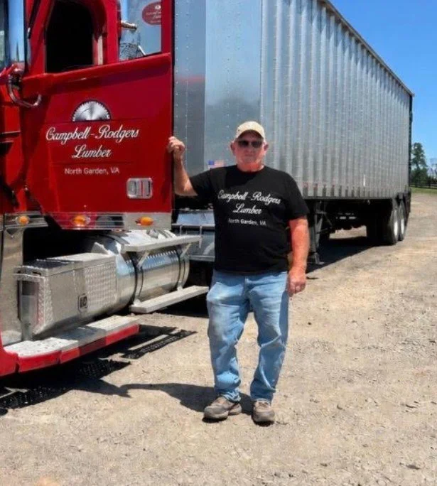 Man standing in front of a red and gray semi-truck, wearing sunglasses, a black T-shirt, and jeans, with clear blue sky in the background.