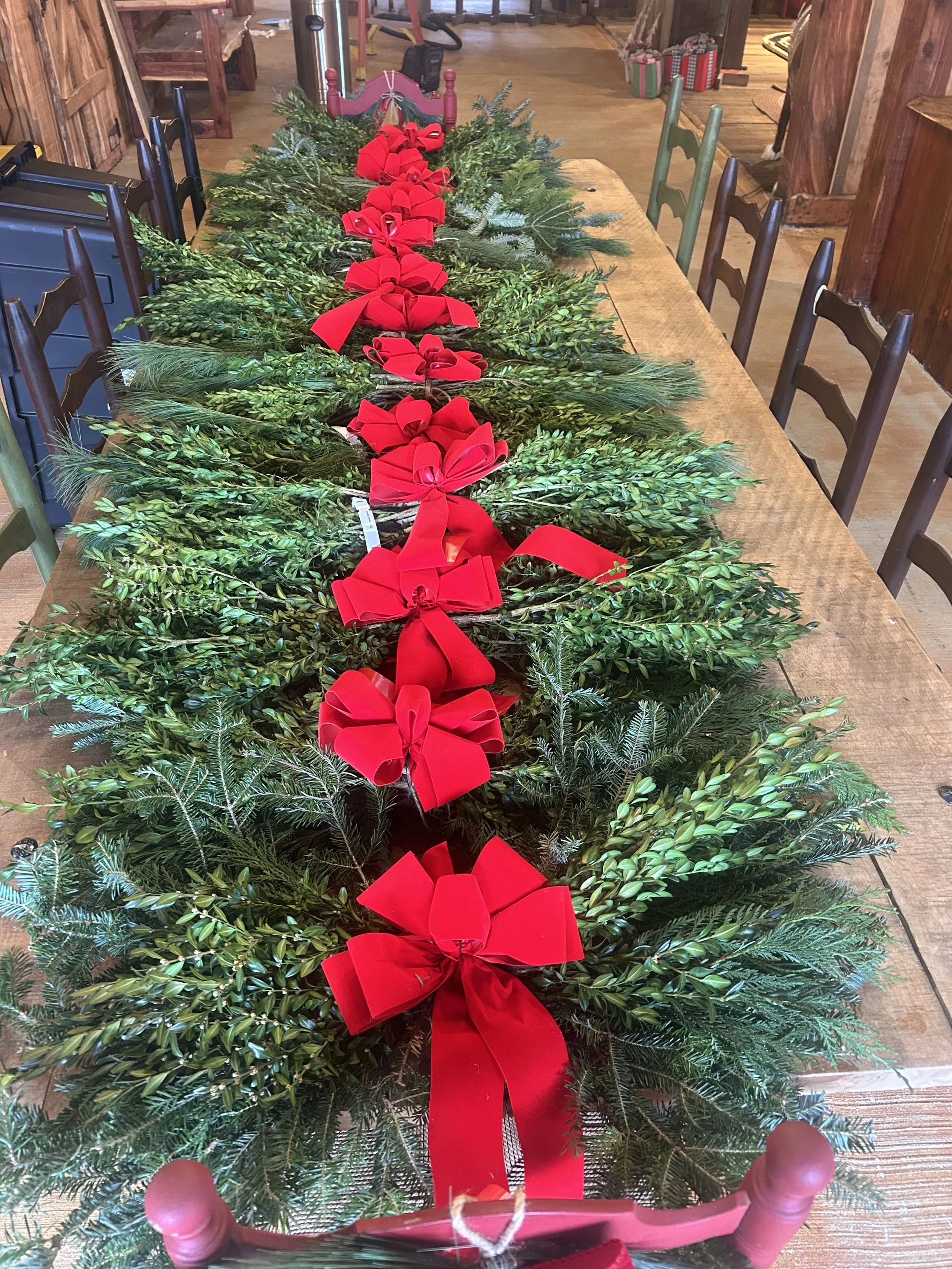 A wooden table holding a long arrangement of green foliage, decorated with red bows and ribbons, with a wooden rocking horse in the foreground and various other furniture and boxes in the background.