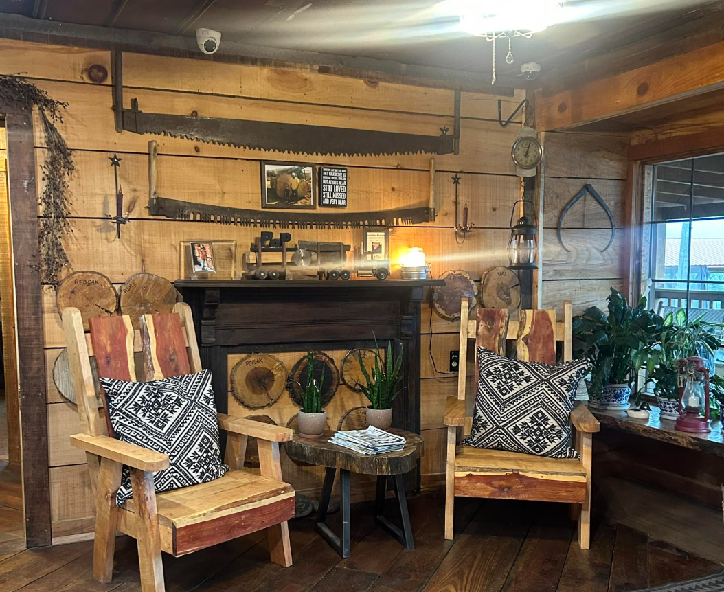 Cozy rustic living room corner with two wooden chairs with patterned cushions, a small coffee table with potted plants and magazines, and a wooden wall decorated with vintage saws, a framed photo, and decorative axe heads. There is a window on the right side with a view outside.
