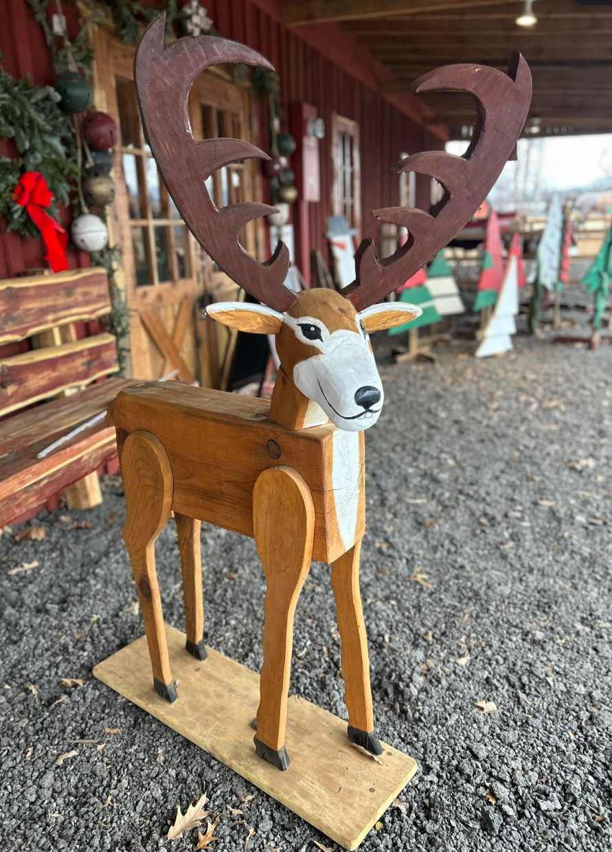 Handcrafted wooden reindeer sculpture with painted face and large antlers, standing outdoors on gravel with a rustic wooden building and Christmas decorations in the background.