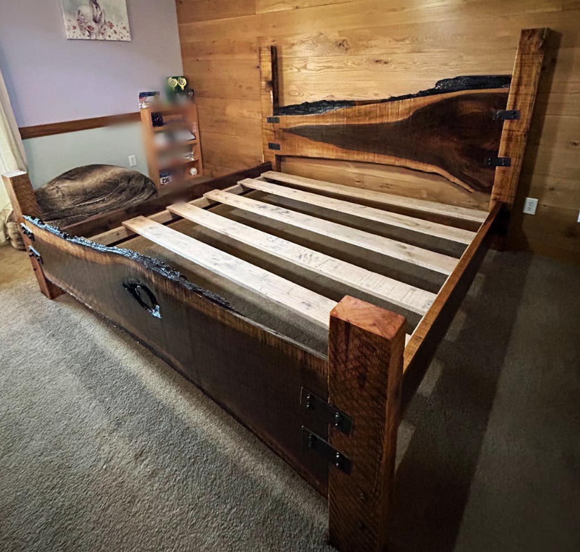 A wooden bed frame with a live edge headboard and side panels, without a mattress, in a bedroom with wood wall paneling and carpeted floor.