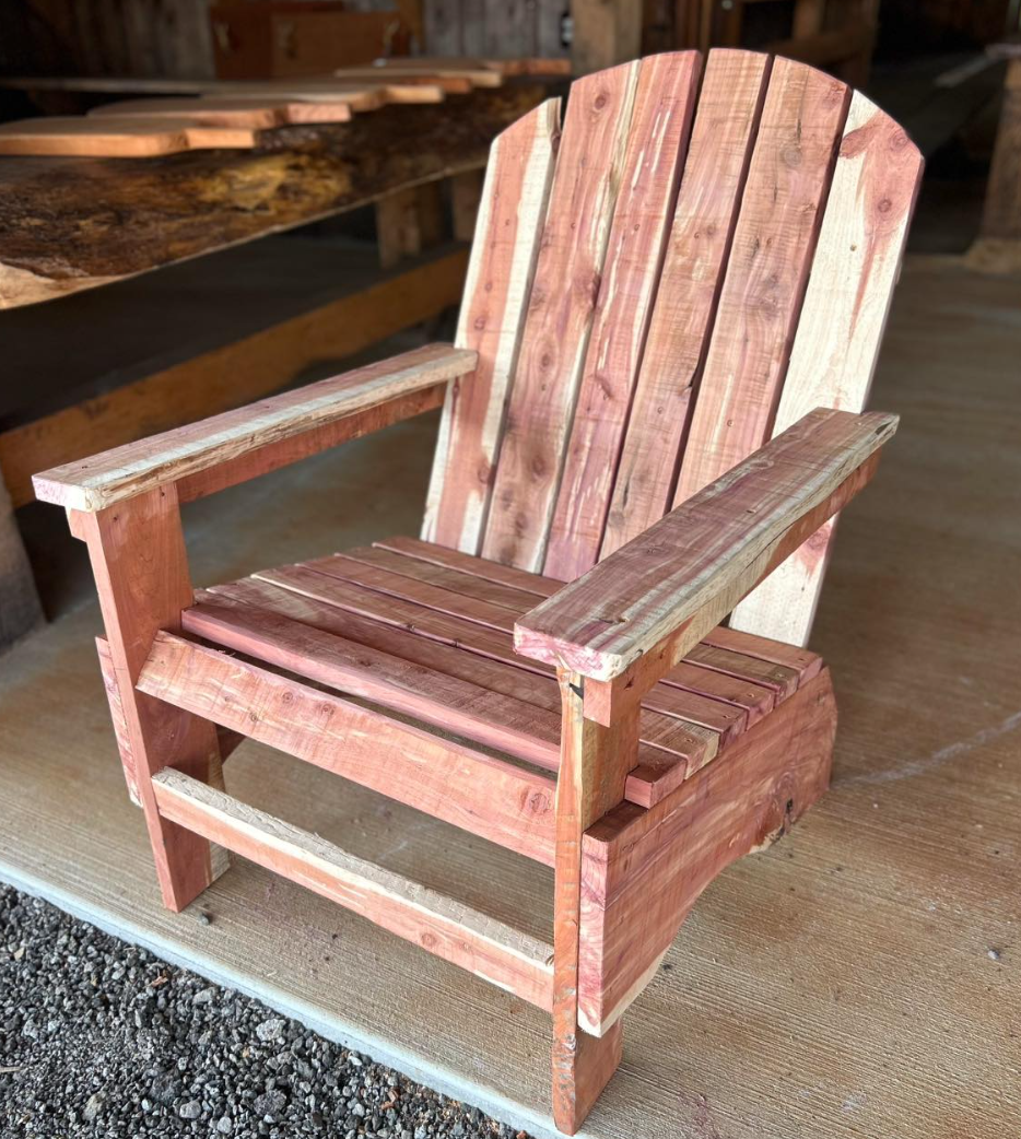 A small Adirondack-style wooden chair made from unfinished or lightly stained wood, placed on a concrete or cement surface with a gravel area in the foreground, and a wooden table or workbench visible in the background.