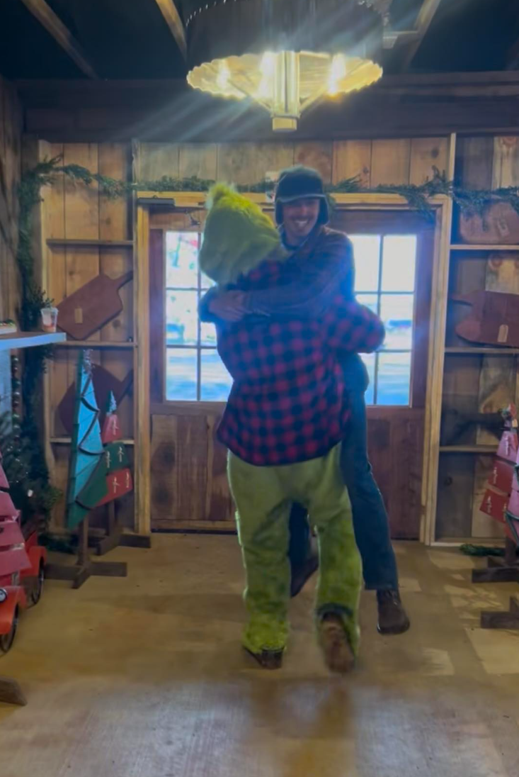 Two people hugging inside a cozy wooden room with Christmas decorations, including tree, ornaments, and gift boxes, near a window with snow outside.