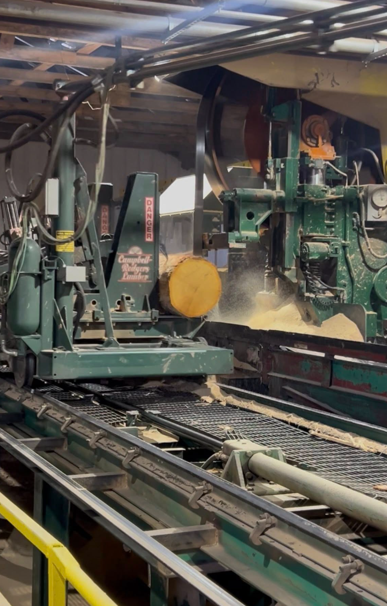 A large industrial machine saws through a log, with sawdust flying as the saw cuts. The machinery is set in a workshop or sawmill environment.