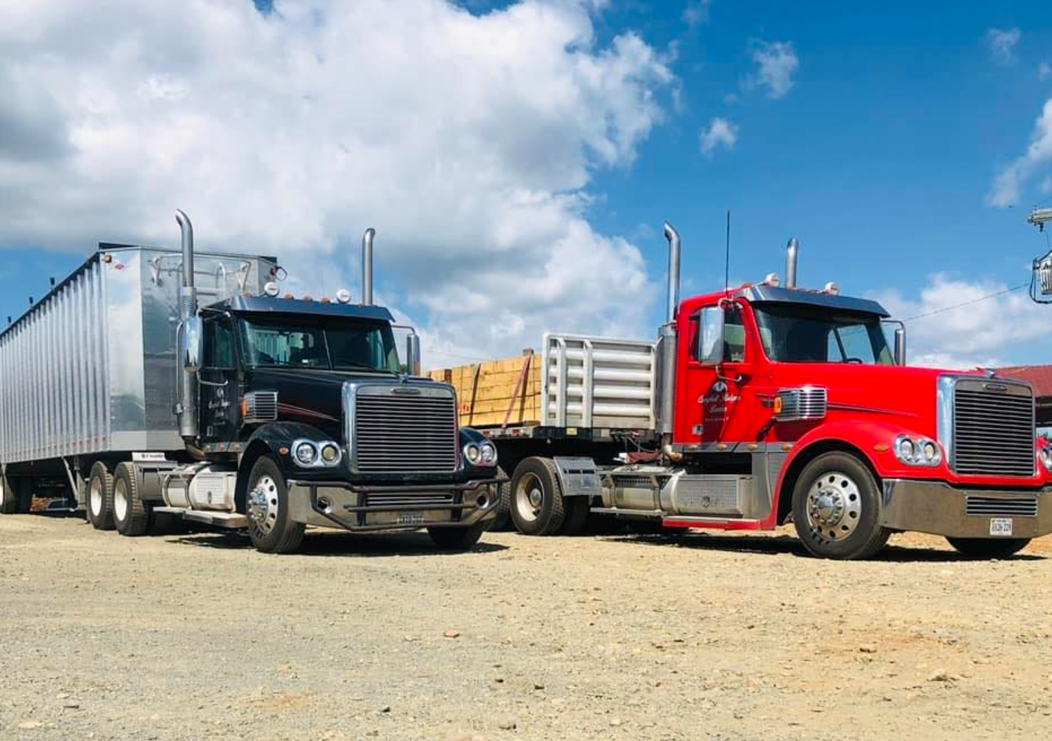 Two semi-trucks parked on a dirt lot under a partly cloudy sky. One truck is black with a silver trailer, and the other is red with a wooden cargo bed.