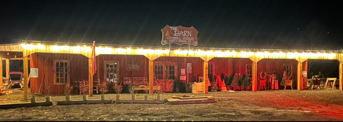 Nighttime view of a rustic wooden barn decorated with Christmas trees and holiday figures, illuminated by string lights.