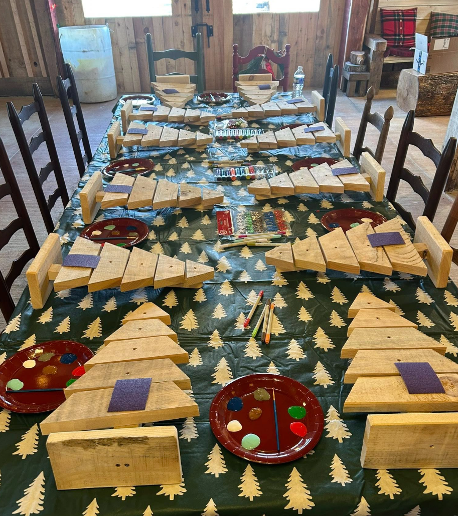 A long table with a festive tree-patterned tablecloth, set with wooden dominoes, colorful paint palettes, and small purple felt pieces, likely prepared for a Christmas-themed activity or game.