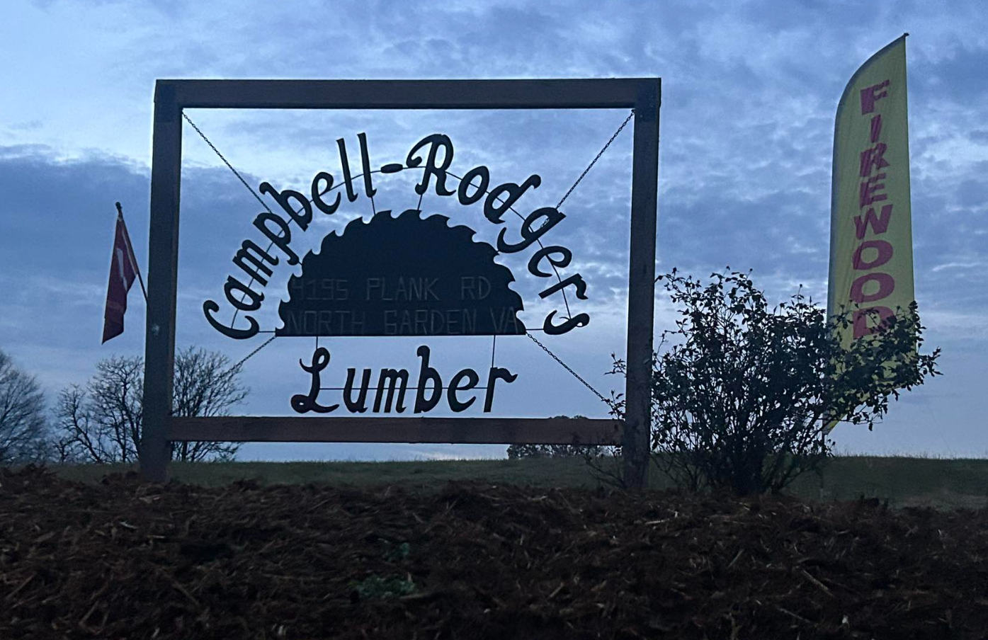 Wooden sign for Campbell-Rogers Lumber with a circular saw blade design in a rural outdoor setting, with trees and flags nearby and a cloudy sky in the background.