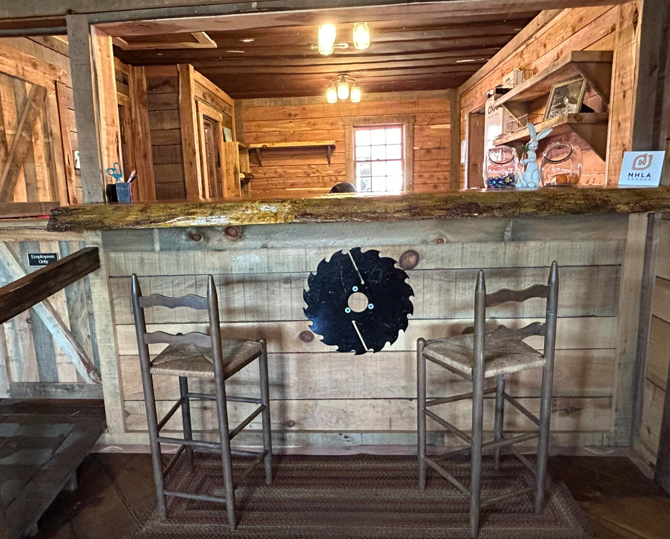 Interior of a rustic wooden bar with two chairs in the foreground, a countertop with a saw blade design on the front, a small shelf with decorative glass bottles and a bunny figurine, wooden walls, and a window letting in natural light.