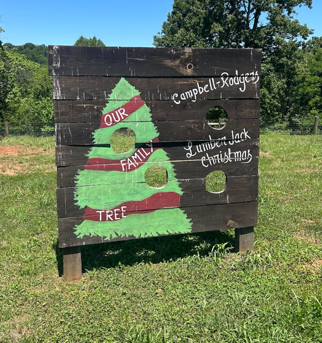 Wooden Christmas tree with cutouts and words, painted with a green tree and red ribbons, on a grassy field with trees in the background.