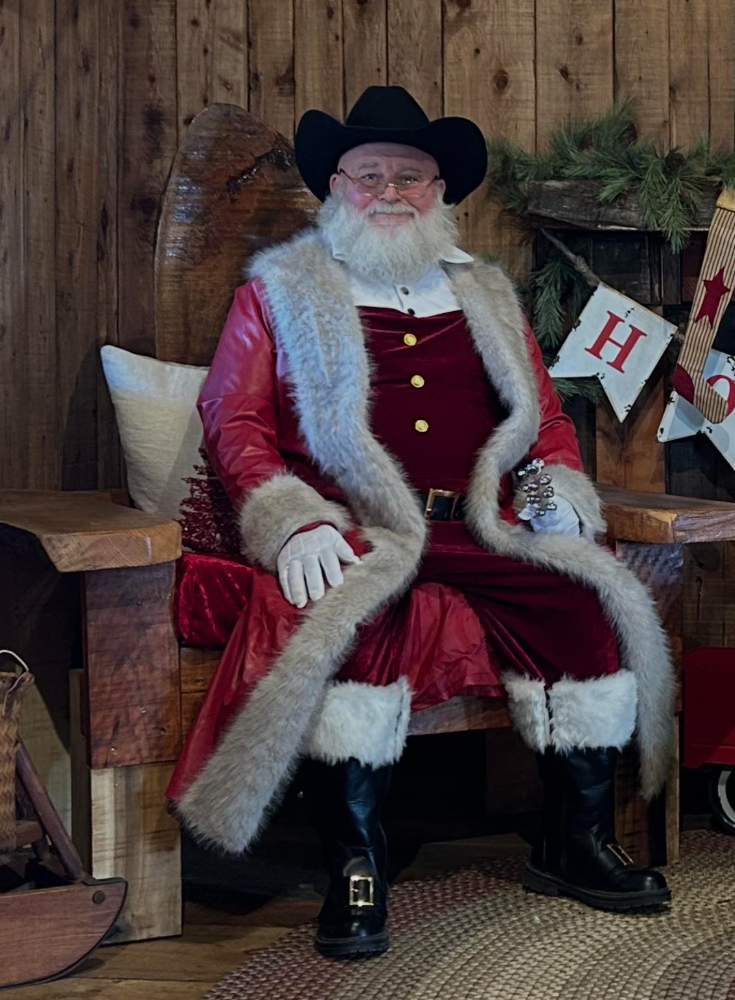 Man dressed as Santa Claus sitting on a wooden bench with a wooden backdrop, wearing a black cowboy hat, red coat with white fur trim, and black boots, with Christmas decorations in the background.