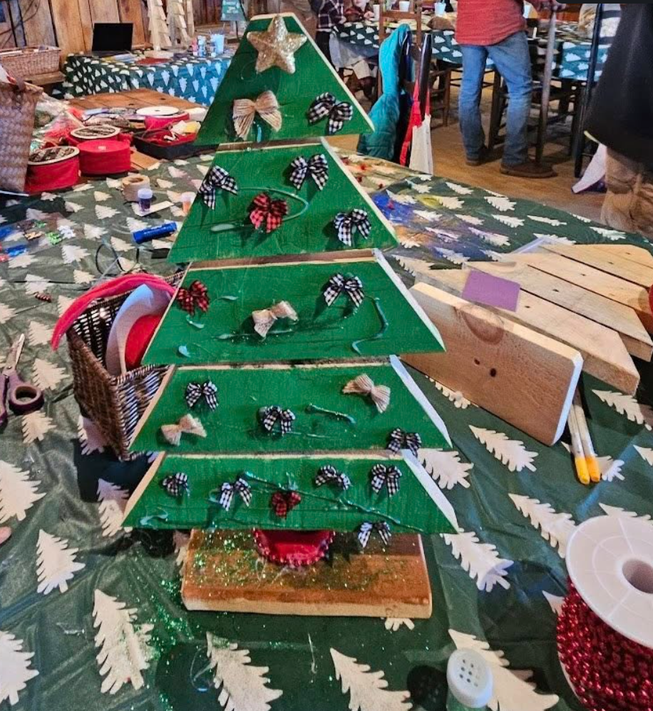 A small, tabletop Christmas tree craft project with three green tiers, decorated with small bows and burlap ribbons, on a festive table with crafting supplies and holiday decorations.